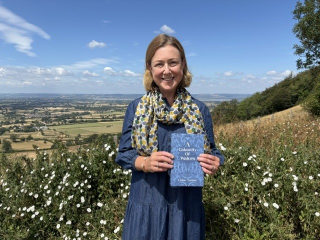 The female author of A Calamity of Waters, Chloe Turner, standing with a copy of the book with her back to the Severn Vale, Gloucestershire. The river is just visible in the distance.